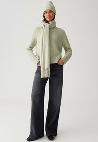 Pull en maille vert clair, bonnet assorti et écharpe légère. Jean large et sombre avec un ourlet effiloché ; bottes noires à bout pointu. Design simple et texturé.