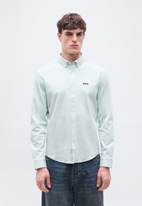 Νεαρός άνδρας που φορά ένα ανοιχτό πράσινο πουκάμισο με κουμπιά και το λογότυπο "BOSS" στο στήθος, καθώς και μπλε τζιν, στέκεται μπροστά σε ένα απλό λευκό φόντο.