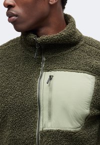 Homme portant une veste polaire vert olive avec un tissu texturé et une poche poitrine zippée lisse, affiché du cou jusqu'au milieu du torse.