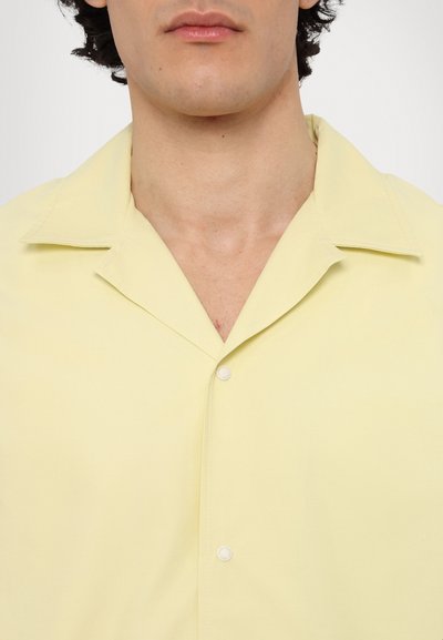 Camisa de botón de manga corta color amarillo claro que presenta un cuello extendido, tela lisa y un solo botón en la parte frontal.