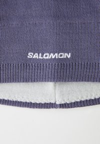 Detalhe de um tecido de lã roxa com forro de lã branca e logotipo bordado em branco "Salomon" na borda canelada.