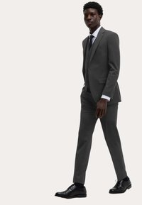 Grå jakkesæt med tre knapper og såkaldte notch-lapel, parret med en hvid skjorte og et mønstret slips. Sorte lædersko fuldender sættet.