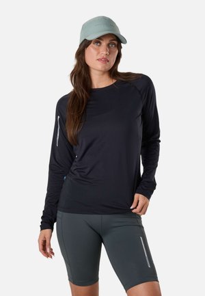 Kvinde iført en lyseblå kasket, sort langærmet skjorte og grå cykelshorts med refleksdetaljer, stående mod en hvid baggrund.