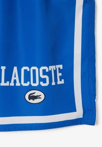 Pantalones cortos deportivos azul con rayas blancas. Presenta un gran texto "LACOSTE" y un logo de cocodrilo negro dentro de un óvalo en la parte frontal.