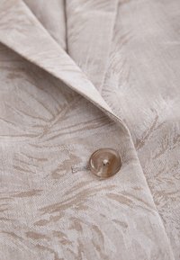 Primer plano de chaqueta beige de tela con patrón sutil de hojas de palma, solapa con muesca y un solo botón redondo translúcido.