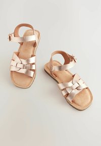 Sandalias metálicas de bronce con tiras trenzadas, hebilla en el tobillo y suela plana. Interior suave similar al ante con un acabado liso.