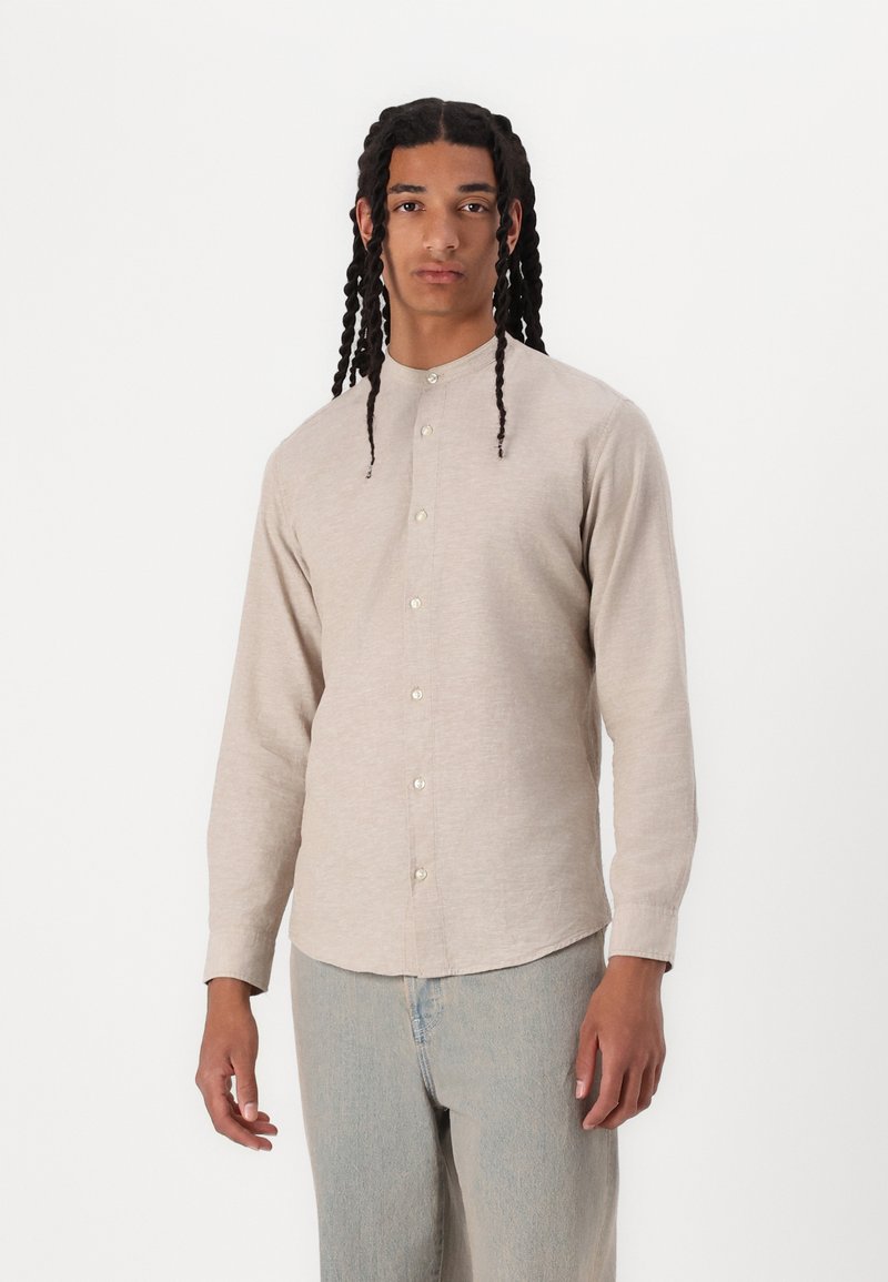 Camicia lunga beige chiaro con colletto alla cinese, chiusura frontale con bottoni e una texture morbida, abbinata a jeans blu chiaro.