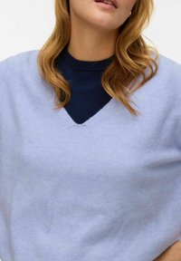 Gaiši zils, mīksti adīts džemperis ar V veida izgriezumu, valkāts virs tumši zilas apļveida kakla krekla. Gluda tekstūra, ikdienišķs piegriezums.