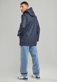 Navy waterproof jacket with a hood, long sleeves, and side pockets. Paired with light blue jeans and white sneakers with black accents.