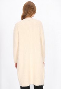 Cardigan lung și lejer, de culoare crem, confecționat dintr-un material tricotat texturat, având mâneci lungi și un tiv rotunjit.