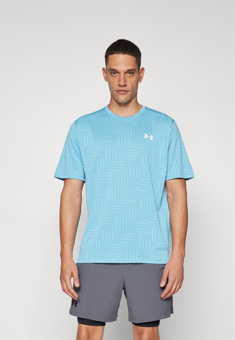 Maglietta sportiva azzurro chiaro con un motivo geometrico, scollatura rotonda e maniche corte. Abbinata a pantaloni corti grigi. Logo Under Armour visibile.