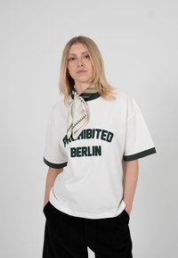 Mlada žena s plavom kosom nosi bijelu majicu na kojoj piše "ZABRANJENO BERLIN", crne hlače i šal s uzorkom vezan oko vrata.