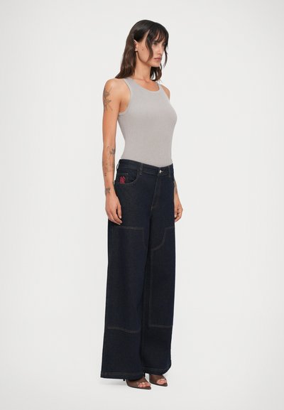 Mulher com tatuagens vestindo uma blusa cinza sem mangas e calças largas escuras, de pé, usando sapatos de salto aberto castanhos, em fundo branco.