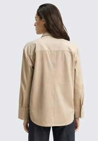 Camisa de manga larga beige con un corte relajado, yoke posterior y dobladillo curvo. Presenta puños abotonados y una textura suave.