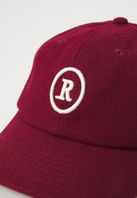 Casquette en laine bourgogne avec un logo circulaire brodé blanc featuring la lettre "R". Sa texture douce et sa visière incurvée sont des caractéristiques de conception notables.