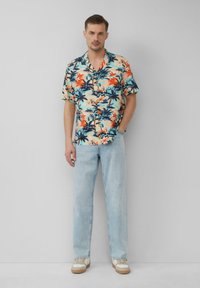 Kurzarm-Hawaiianhemd mit einem Blumen- und Palmenmuster in Blau, Orange und Creme; kombiniert mit hellblauen Jeans und Sneakers.