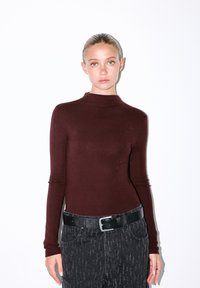 Bordeaux langemouwen top met een hoge hals, gecombineerd met gestructureerde zwarte jeans en een platte, zwarte riem. Glad materiaal met subtiele patronen.