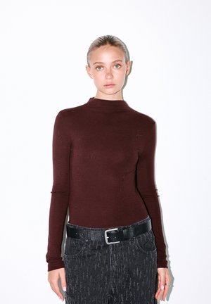 Top lung cu mâneci lungi de culoare burgundy, cu guler înalt, asortat cu blugi negri texturat și o curea plată neagră. Material neted cu modele subtile.