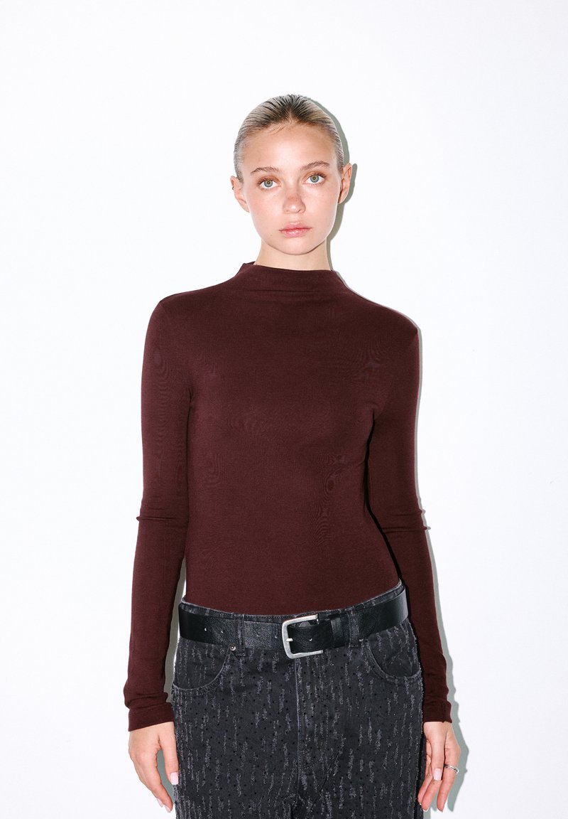 Bordeaux langemouwen top met een hoge hals, gecombineerd met gestructureerde zwarte jeans en een platte, zwarte riem. Glad materiaal met subtiele patronen.