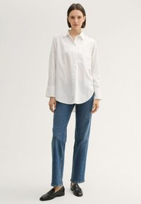 Weißes Hemd mit Kragen und Vordertasche, kombiniert mit blauen geraden Jeans und schwarzen Slippern, vor einem neutralen Hintergrund.