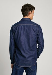 Donkerblauwe denim overhemd met lange mouwen, een kraaghals, zichtbare stiksel-details en knoopmanchetten. Rechte zoom met subtiele textuur.