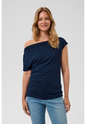 Ung kvinde med langt blond hår iført en navyblå top med én skulder og lyseblå jeans, står og smiler foran en ensfarvet baggrund.
