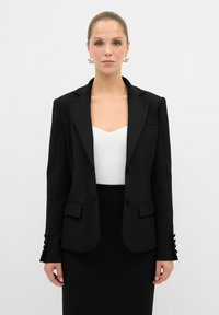 Blazer nero su misura con rever a punta, due tasche frontali e bottoni decorativi sulle maniche. Indossato sopra un top bianco e una gonna nera.