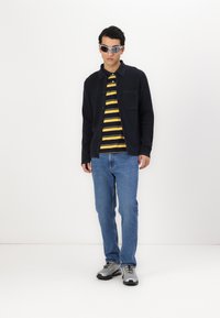 Melna cardigan, dzeltena un melna svītraina polo krekls, zila džinsu bikses un pelēki sporta apavi. Modelis valkā sudraba saulesbrilles un stāv uz balta fona.