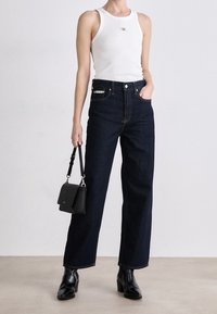 Dunkelblaue, hochgeschnittene Jeans mit weitem Bein, kombiniert mit einem taillierten weißen Tanktop und einer kleinen schwarzen Handtasche. Glatte Denim-Oberfläche, minimaler Hardware.