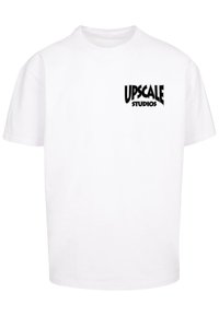 Biela bavlnená tričko s čiernym logom "UPSCALE STUDIOS" na hornej ľavej strane, voľný strih s krátkymi rukávmi.