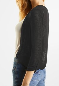 Svart, lättvikts, öppen cardigan med trekvartslånga ärmar, med en texturerad design och avslappnad passform, kombinerad med en beige top och blå jeans.