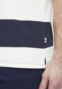 Bavlnená polo košeľa s horizontálnymi námorníckymi modrými a bielymi pruhmi. Obsahuje bočné ventily a malý značkový akcent v červených, bielych a modrých farbách.