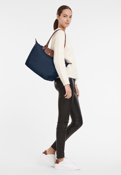 Navy blue tote bag made of durable material, featuring a brown leather handle and a gold logo detail. Bag has a spacious, structured design.