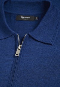 Pulovr z merino vlny v námořnické modři s límcem na zip. Má hladkou texturu a kovový zip s malým logem. Viditelná velikost L.