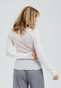 Langärmliges, weißes Hemd aus leichtem Stoff mit etwas transparenter Textur. Bietet eine lockere Passform und Rüschenmanschetten. Kombiniert mit grauen Hosen.