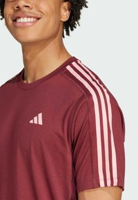 Camiseta deportiva color granate con diseño de tres franjas rosas en los hombros y un pequeño logo rosa en el pecho. Tejido liso y ligero.