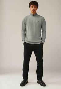 Pull en maille gris avec un col rond, associé à un pantalon noir et des chaussures noires. Présente un petit logo brodé sur le pull.