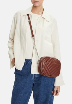 Mujer con chaqueta color crema y vaqueros azules, llevando un pequeño bolso bandolera marrón acolchado con cadena y tira de cuero.