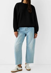 Svart överdimensionerad sweatshirt, ljusblå vida jeans med synliga sömmar och vita sneakers med svarta accenter och gummisulor.