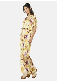 Rumeno in vijolično batik jumpsuit z ohlapnimi rokavi in elastičnim pasom, izdelan iz lahkotnega materiala. Uporablja se z bež sandali na peto.