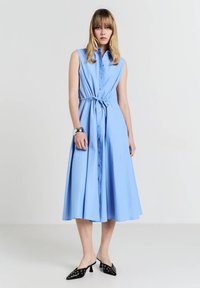 Himmelblaues, ärmelloses Knopfkleid mit einem Gürtel in der Taille, fließendem Rock und glatter Textur. Getragen mit schwarzen Mules mit dekorativen Akzenten.