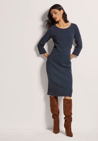 Marineblaues Kleid mit dreiviertel Ärmeln, runder Halslinie und vertikalen Nähten. Kombiniert mit braunen Wildleder-Stiefeln bis zum Oberschenkel.