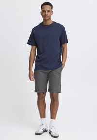 Camiseta de algodón azul oscuro combinada con pantalones cortos gris claro, con los puños doblados y calcetines blancos. El modelo lleva zapatillas deportivas grises y blancas.