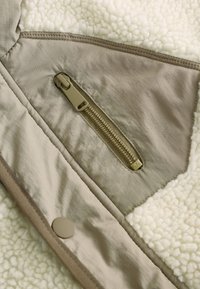 Gros plan sur une veste beige avec doublure en polaire, montrant une poche zippée et un bouton-pression sur un tissu texturé.