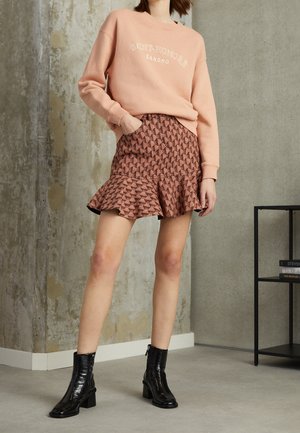 Femme portant un sweat-shirt pêche avec le texte "SAINT-HONORE SANDRO", une mini-jupe marron à volants et motifs, ainsi que des bottines à talons noires en intérieur.