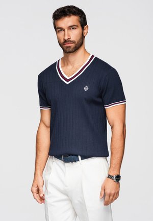 Man met baard, gekleed in een marineblauw geribd V-hals shirt met witte en rode bies, witte broek, marineblauwe riem en zilveren polshorloge, staand tegen een grijze achtergrond.