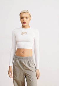 Baltas, ilgas rankoves trumpas marškinėliai su auksiniu "Nike" logotipu priekyje; derinami su šviesiai pilkomis elastinėmis kelnėmis, lygiu paviršiumi, sportiniu pritaikymu.