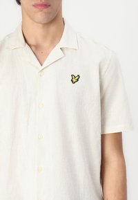 Camicia a maniche corte di colore crema con texture in lino. Presenta un logo di un uccello ricamato in nero e giallo sul petto e chiusura con bottoni.