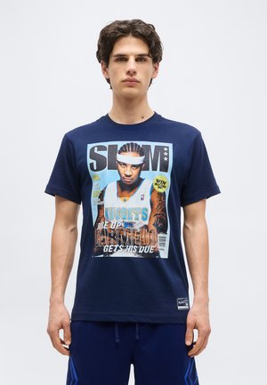 Młody mężczyzna w granatowej koszulce z kolorową okładką magazynu o koszykówce, na której na przedzie widnieje zawodnik Carmelo Anthony.