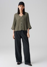 Camicia button-up verde oliva, con texture, col v a maniche a sbuffo, abbinata a pantaloni neri larghi e décolleté neri.